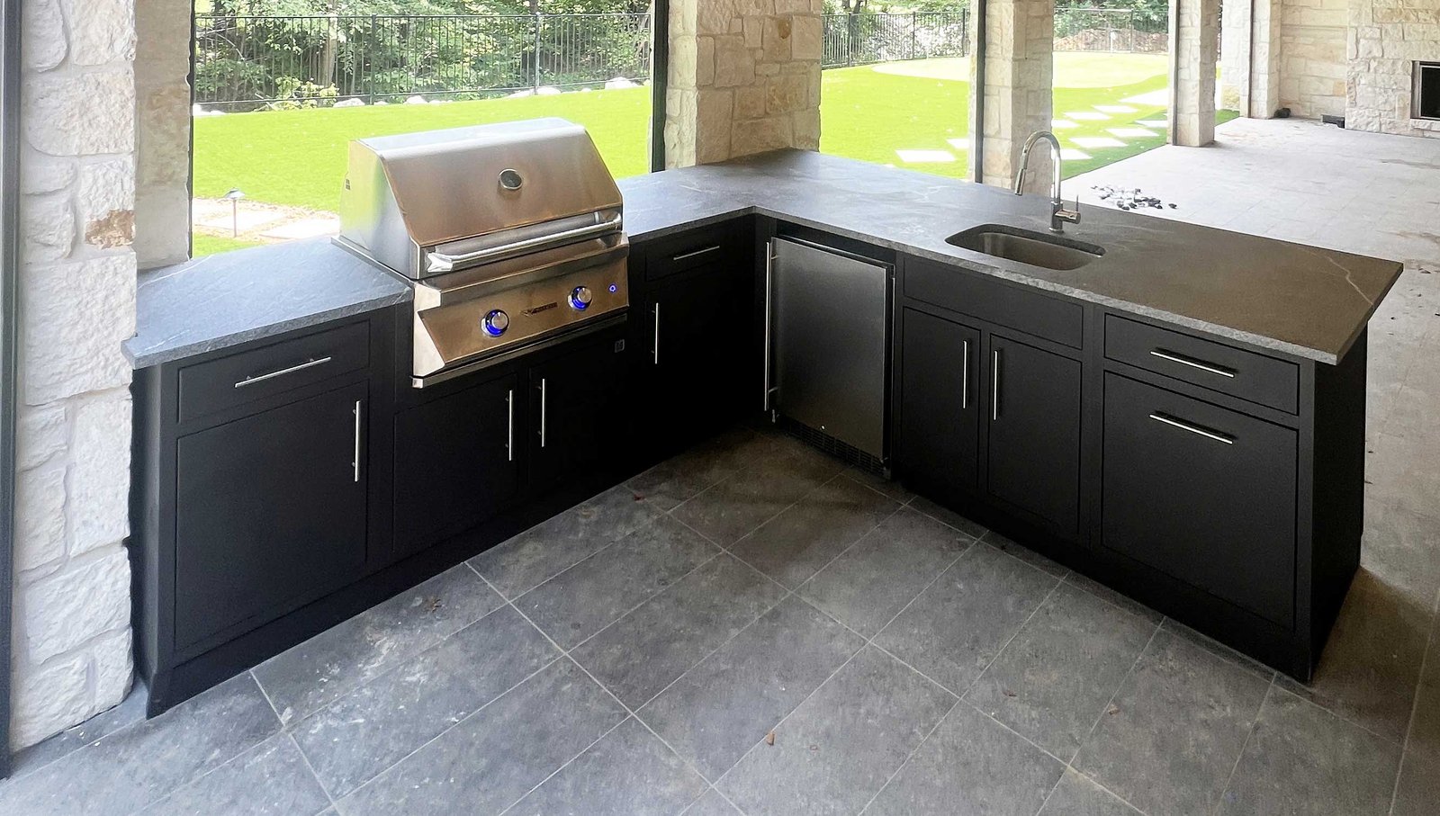 Graphite-finish outdoor kitchen island with a built-in grill, refrigerator, and sink arranged in an L-shape under a covered patio